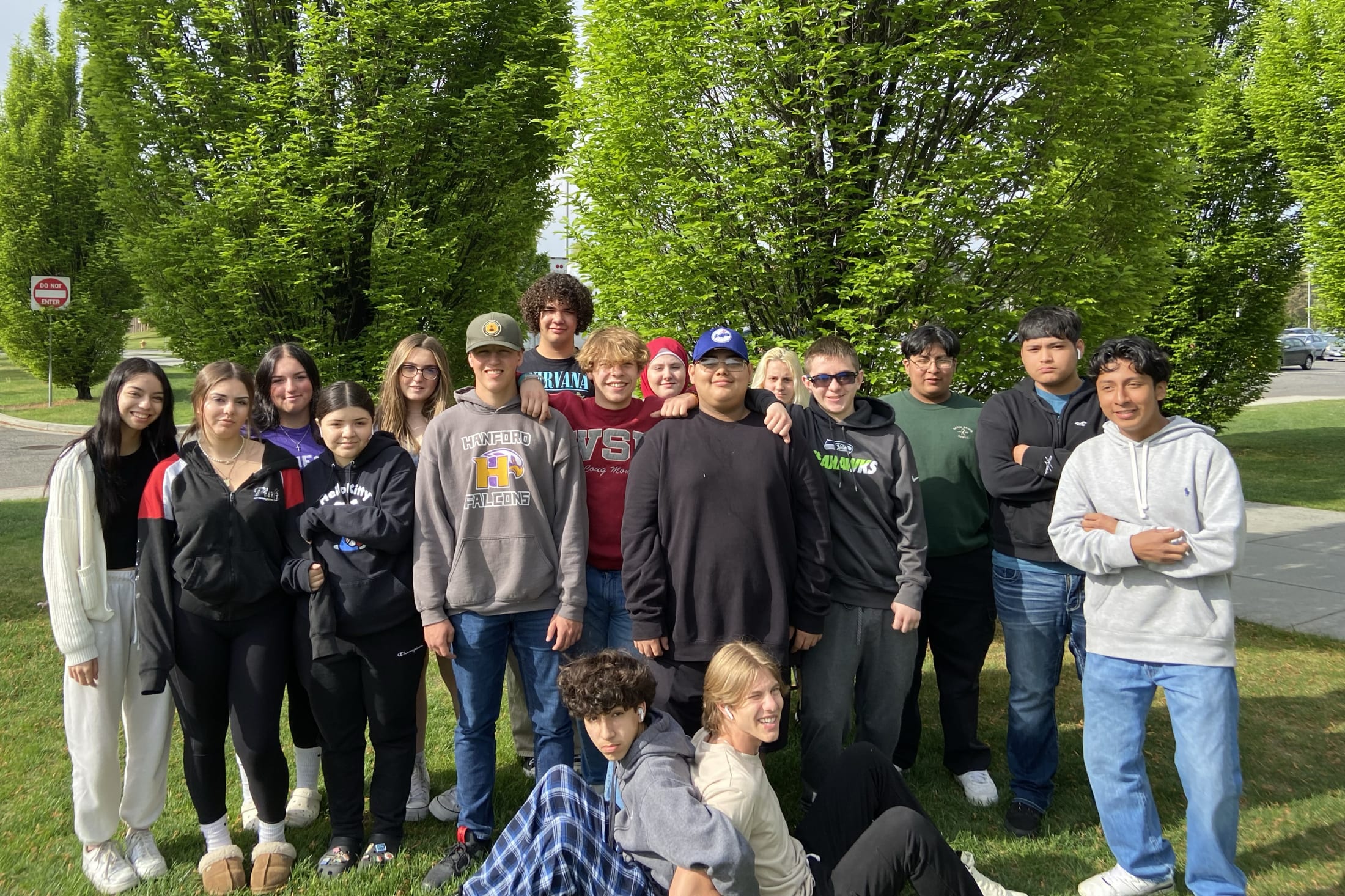 A group of young people, both male and female, standing together in a park-like setting with lush green trees and foliage in the background.