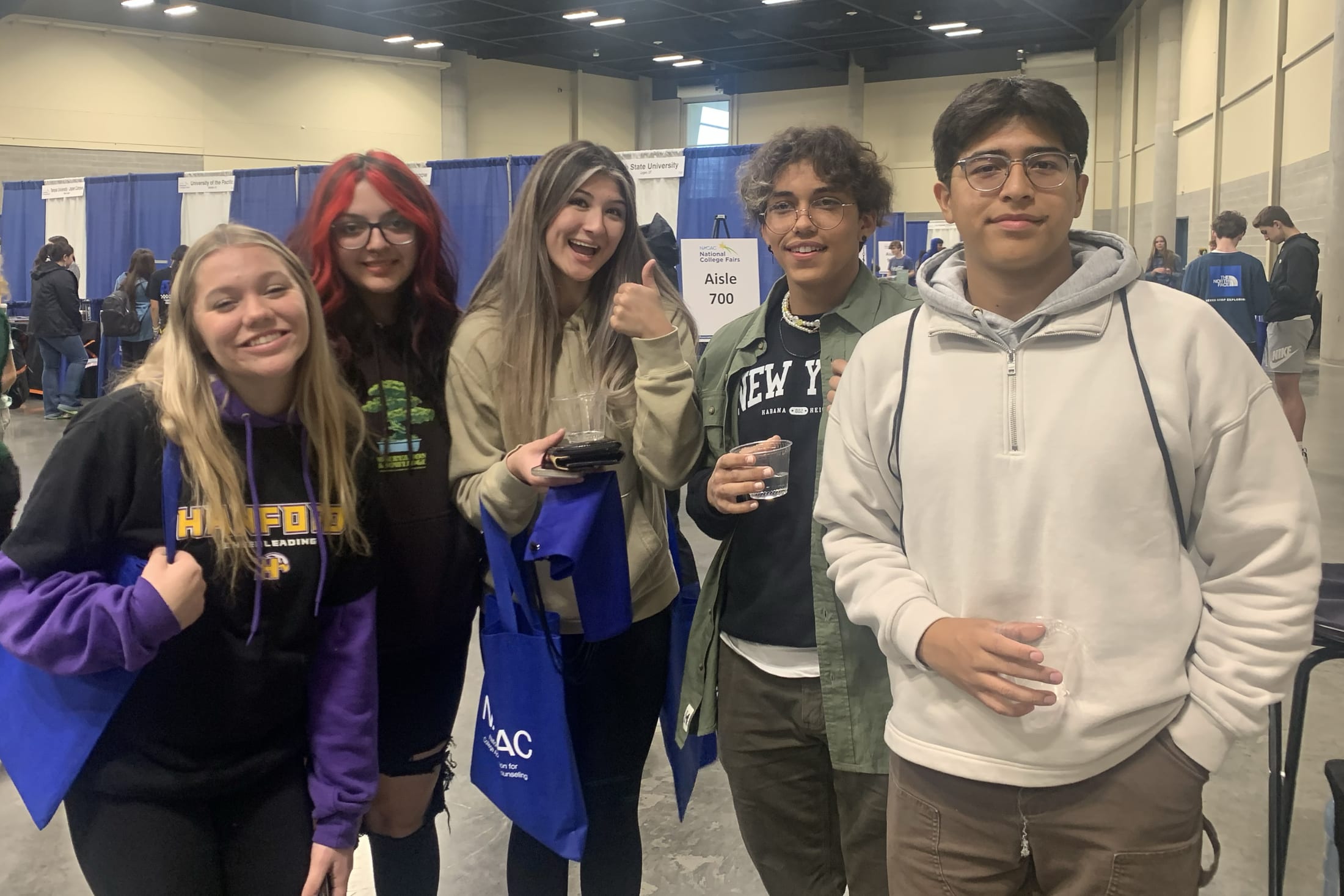 A group of young people, some wearing casual clothing and others in hoodies, are standing together in what appears to be a convention or event space with blue partitions in the background.