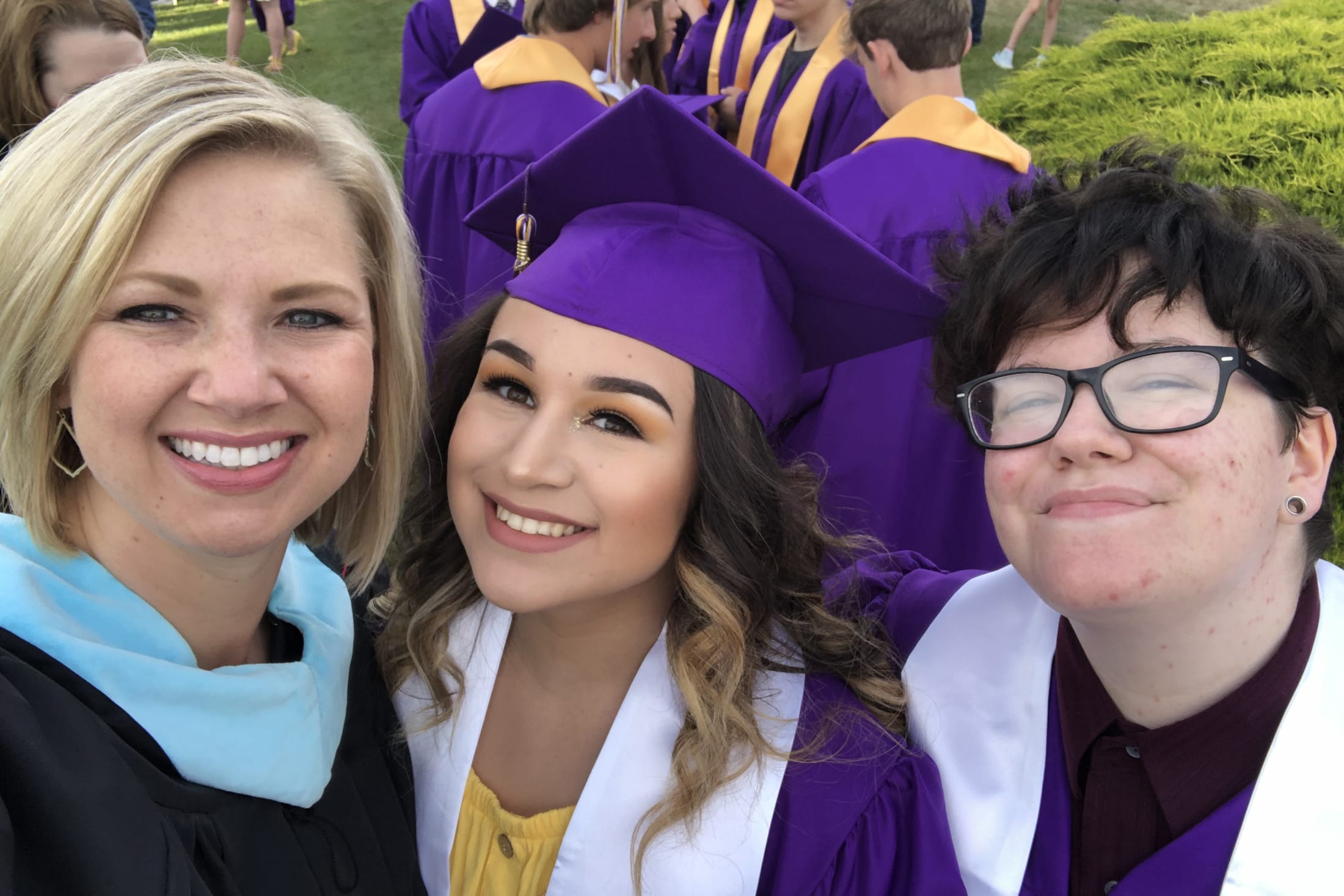 The image shows three people in graduation gowns and caps, with one person in the foreground wearing a blue scarf and the other two wearing purple gowns. They appear to be posing for a photograph in an outdoor setting with other graduates in the background.