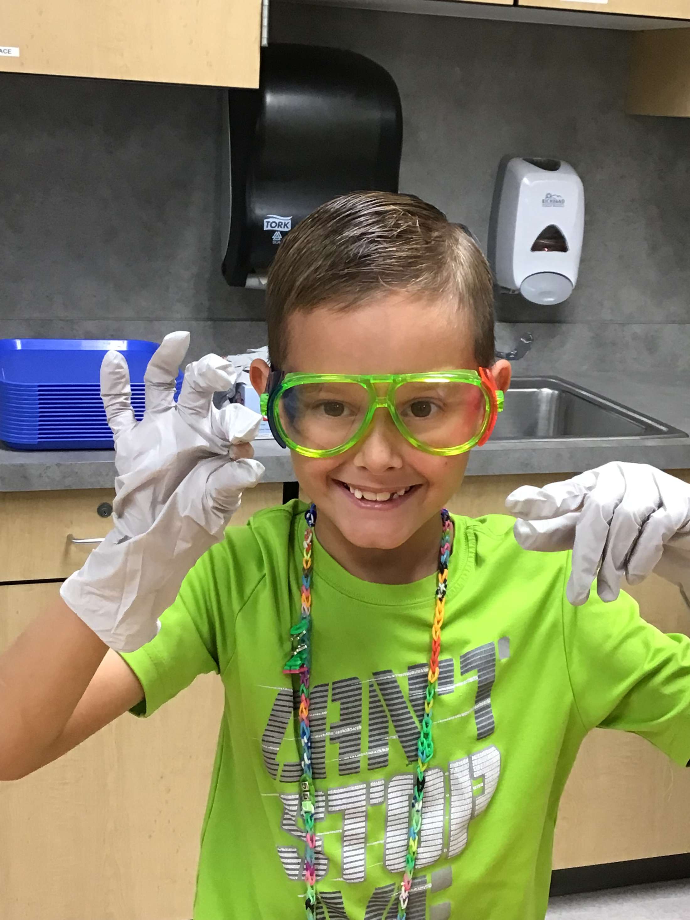 A young child wearing bright green clothing and large, colorful glasses is smiling while standing in a kitchen environment with various appliances and equipment visible in the background.