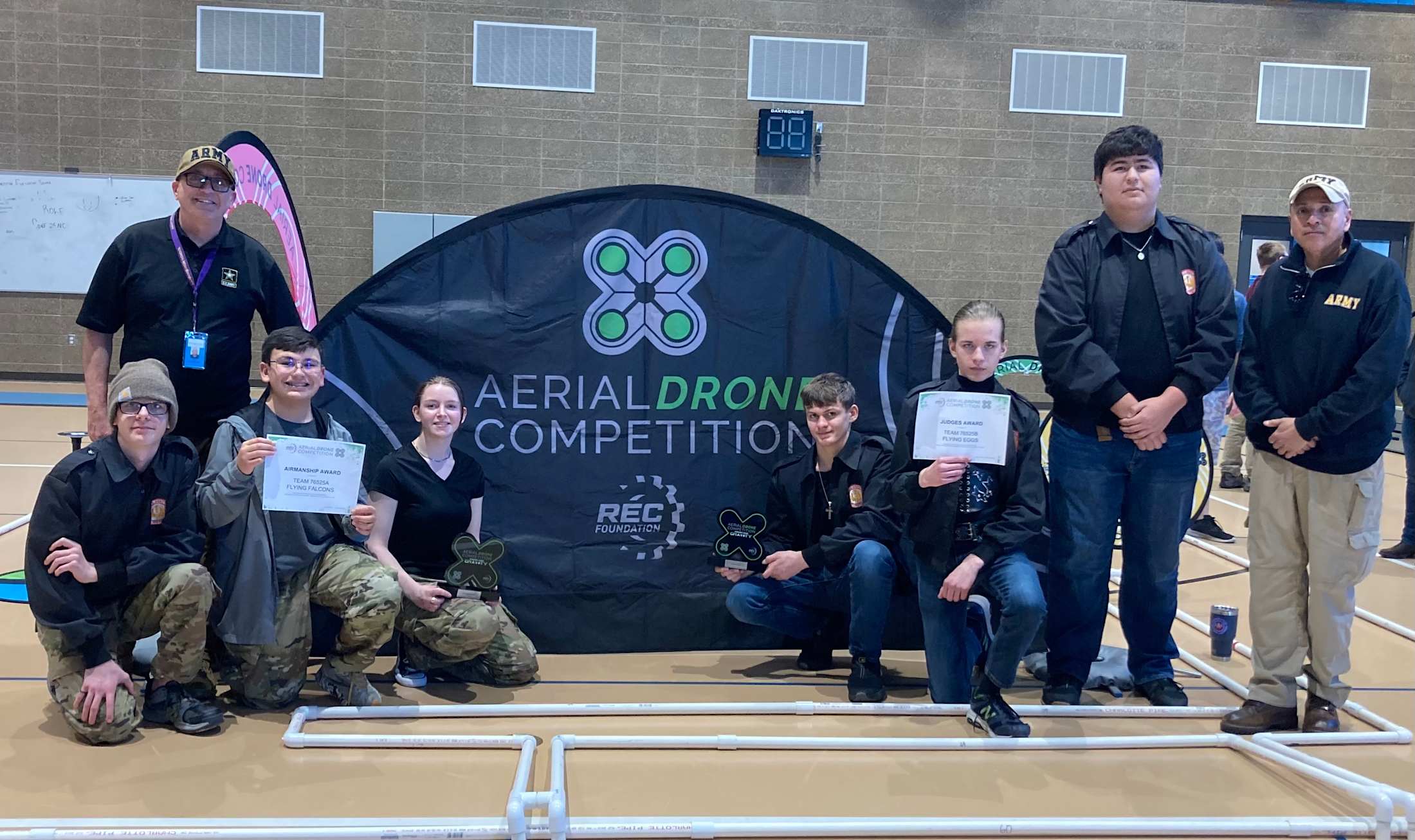 The image shows a group of people posing in front of a large banner for an %22Aerial Drone Competition%22, with several individuals holding certificates or awards.