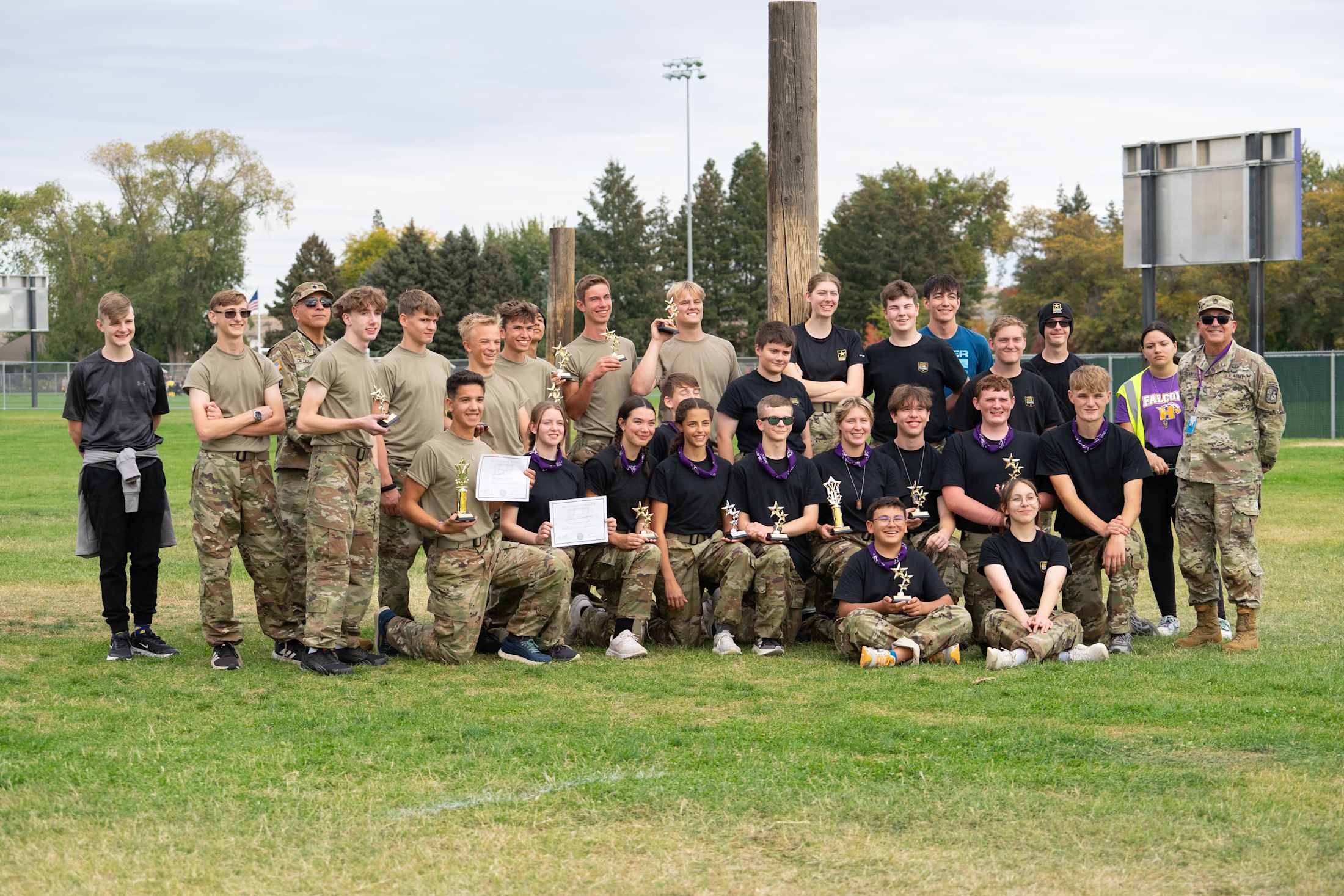 A large group of people, both men and women, are gathered on a grassy field, some wearing military-style uniforms and others in casual attire. In the background, there are trees and buildings, suggesting an outdoor setting.