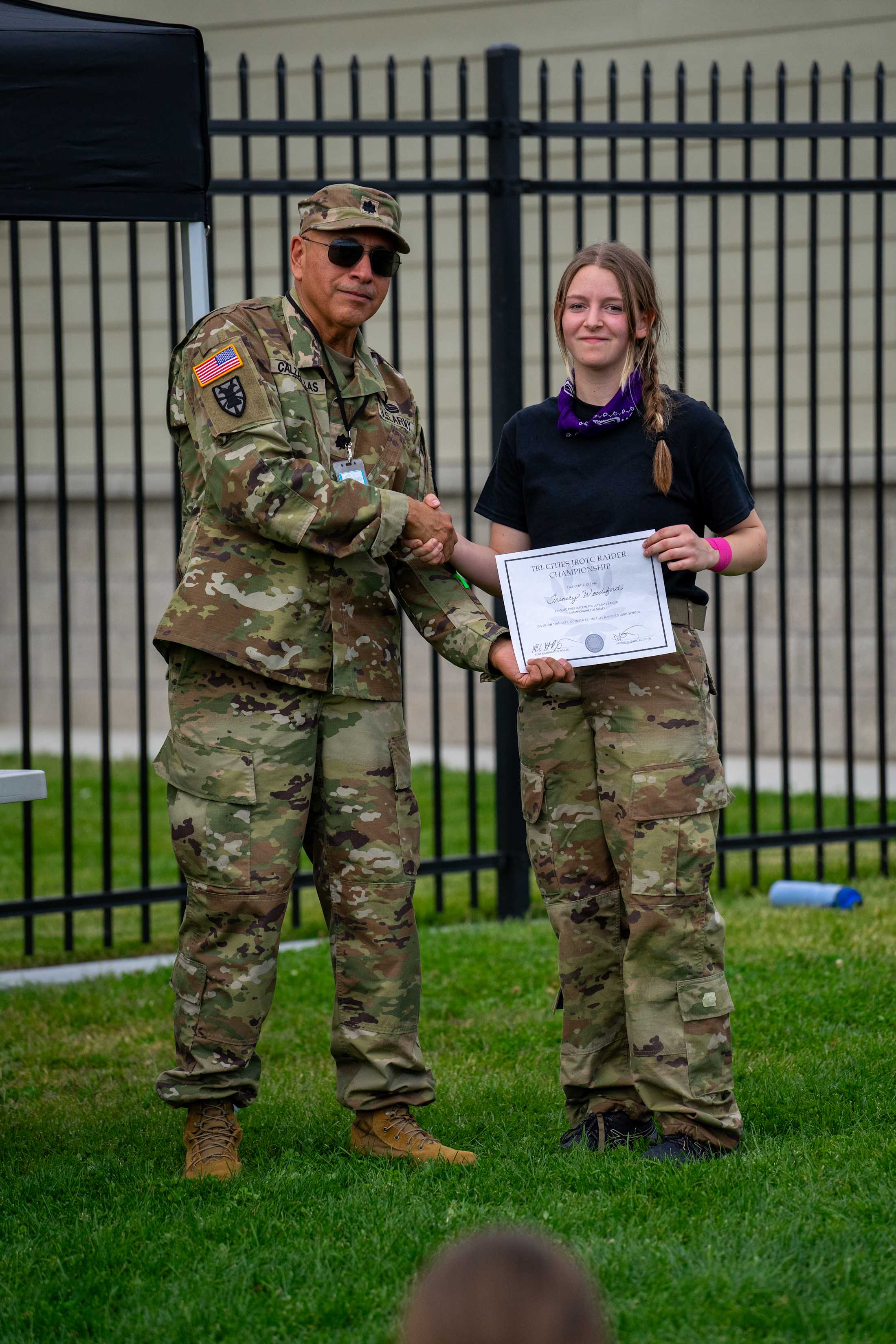 Two individuals in military uniforms, one presenting a certificate to the other, standing in front of a fenced area with a grassy background.