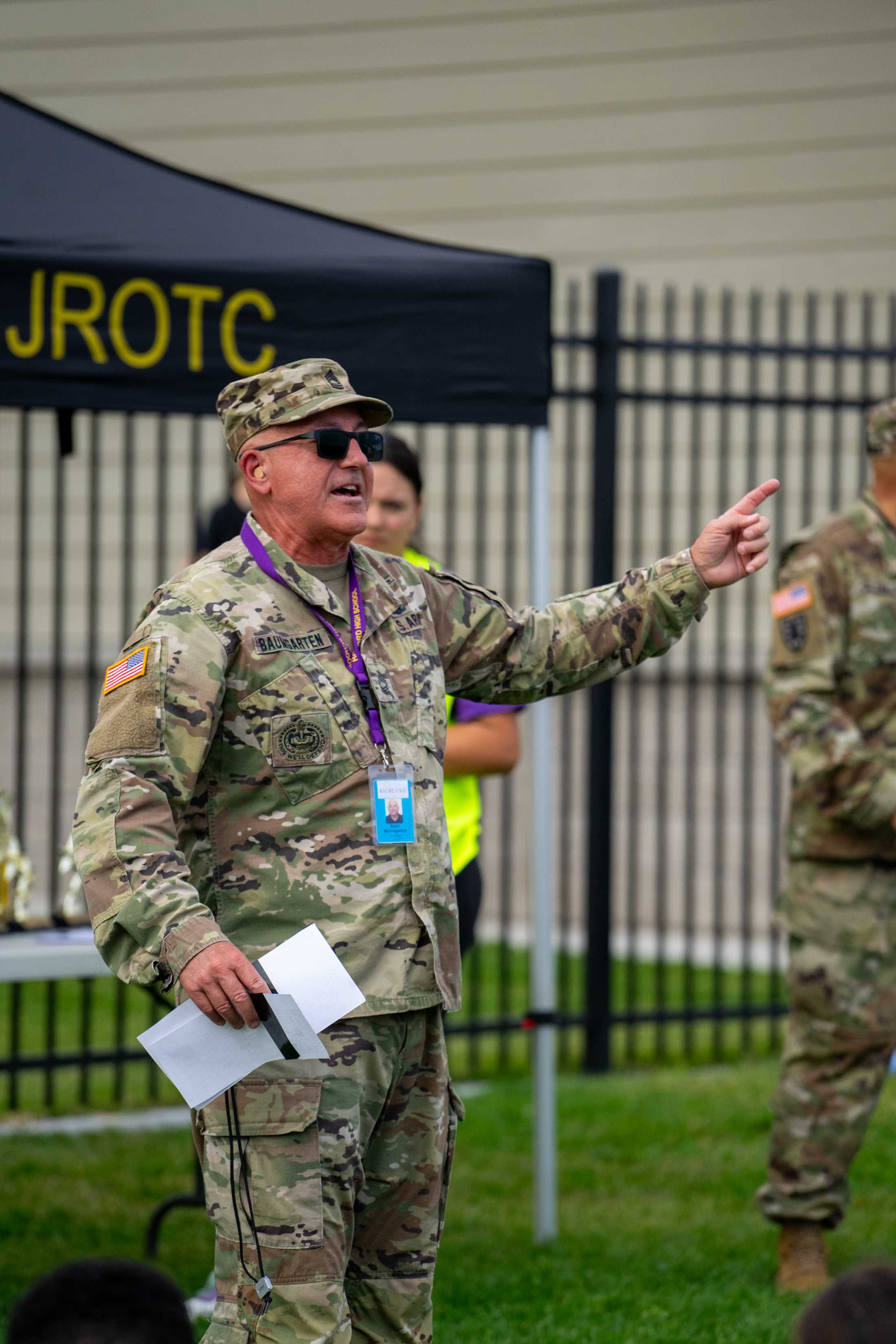 A military officer in camouflage uniform stands in the foreground, holding a document and gesturing with his hand, while a JROTC sign is visible in the background.