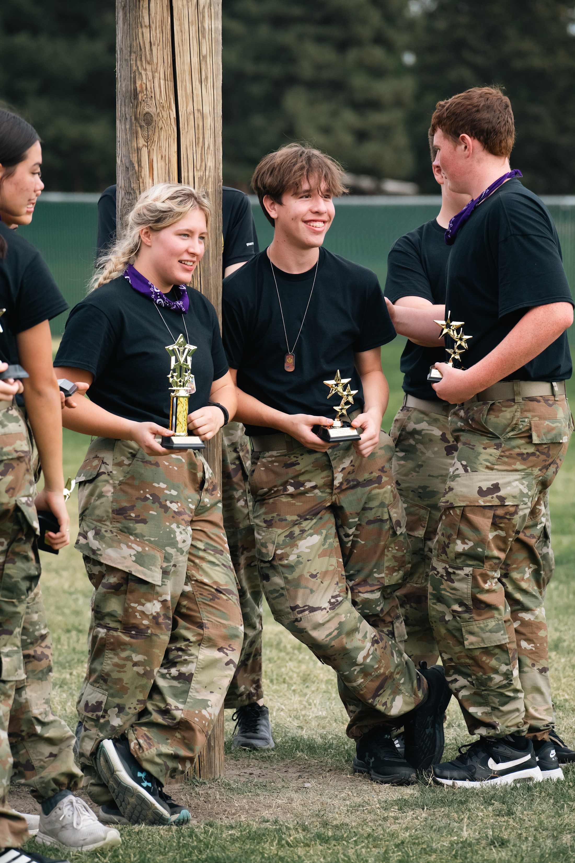 A group of people in military-style uniforms and camouflage pants are gathered outdoors, holding trophies or awards.