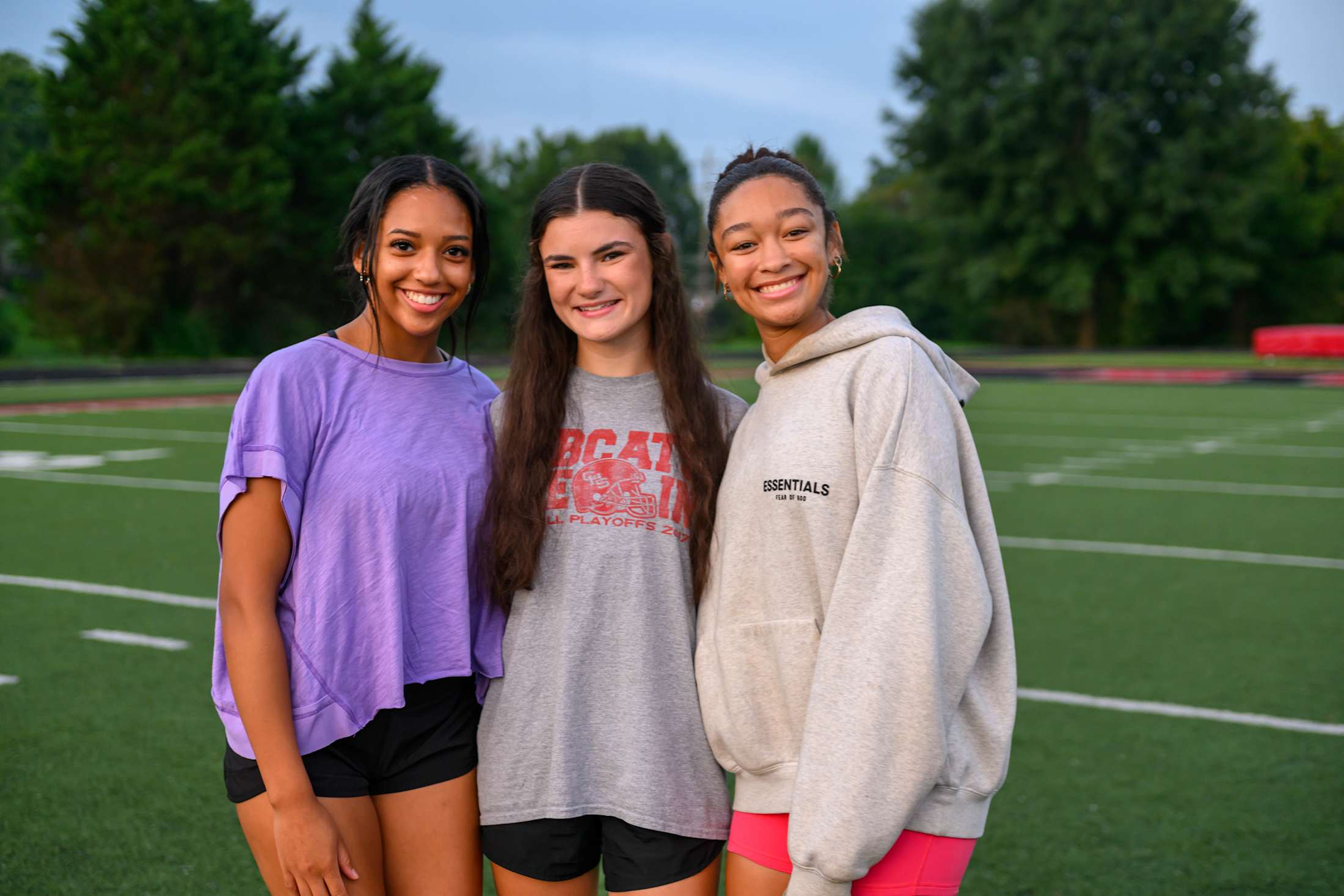 Three young women with long dark hair are standing together on a sports field, surrounded by trees in the background.