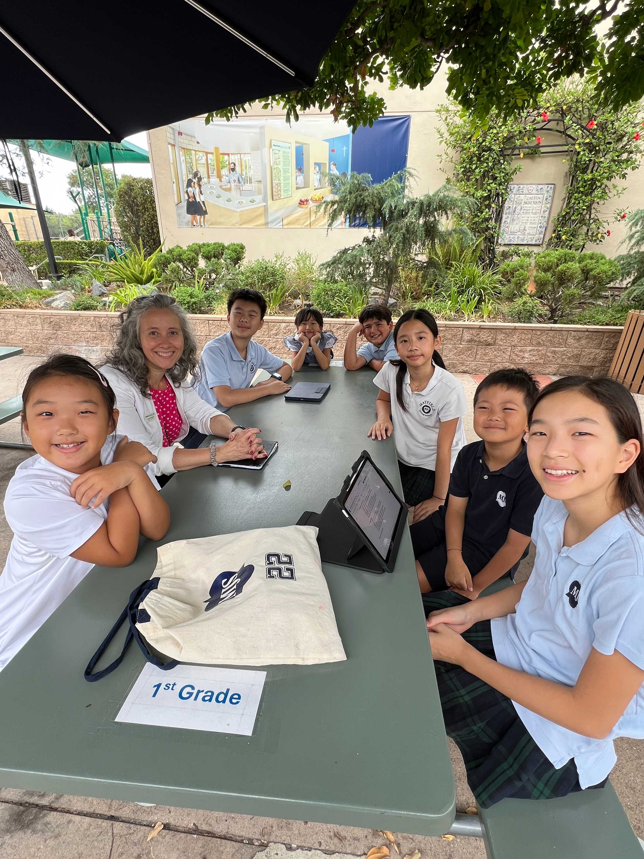 Eight stduents of various ages sitting around a outdoor linch table in a meeting and smiling at the camera.