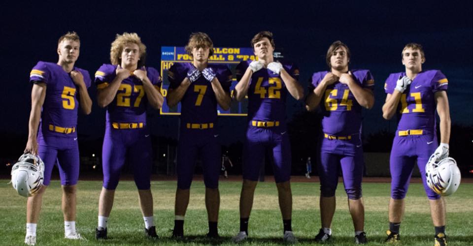 A group of football players in purple uniforms standing together on a football field with a scoreboard visible in the background.
