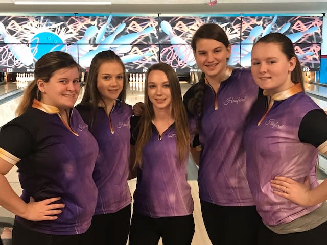 The image shows a group of five young women wearing matching purple bowling shirts, posing together in what appears to be a bowling alley with a colorful, abstract mural on the wall behind them.