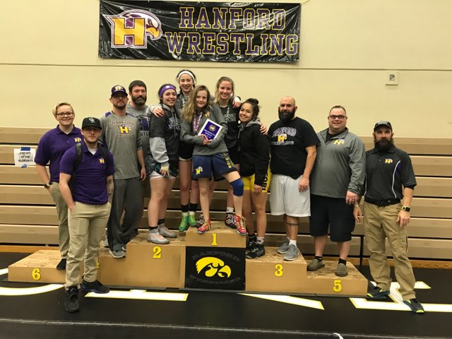 A group of people, some wearing wrestling uniforms, stand on a podium with a %22Hanford Wrestling%22 banner in the background.
