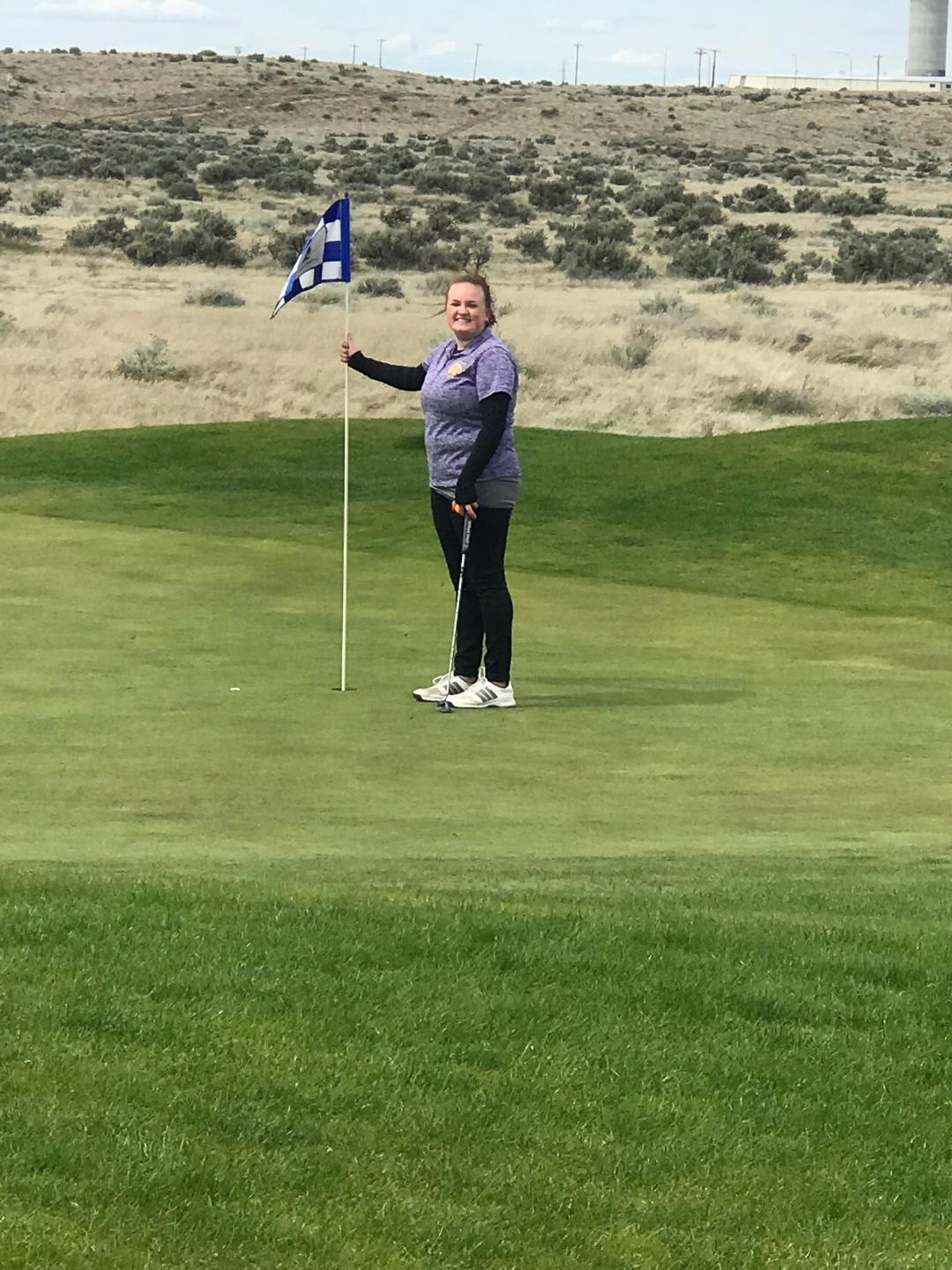 A person in a blue and gray outfit is standing on a well-manicured golf green, holding a flag in the middle of a vast, arid landscape.