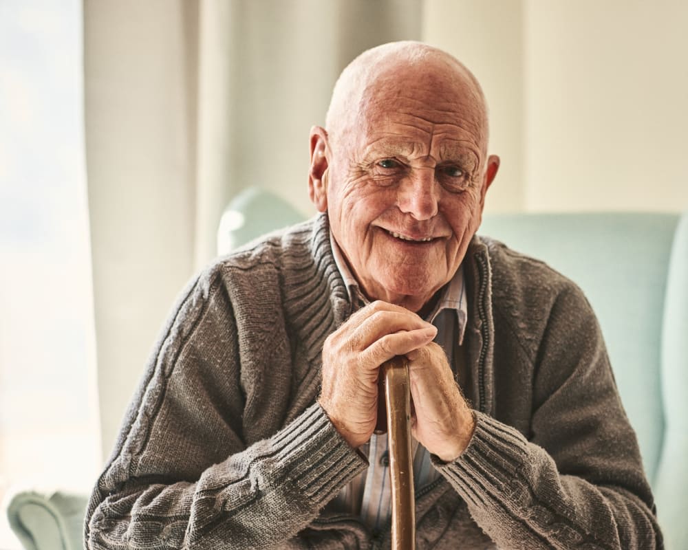 A smiling elderly man with a bald head and wrinkled face, wearing a gray sweater, sits in a chair against a light-colored background.