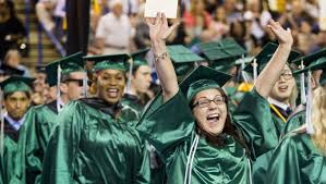 A large group of graduates in green gowns and caps enthusiastically celebrating their accomplishment, with a blurred background of other graduates.