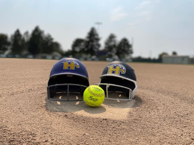 Two baseball helmets with the letter %22H%22 on them, along with a yellow softball, sit on a dirt baseball field with trees in the background.