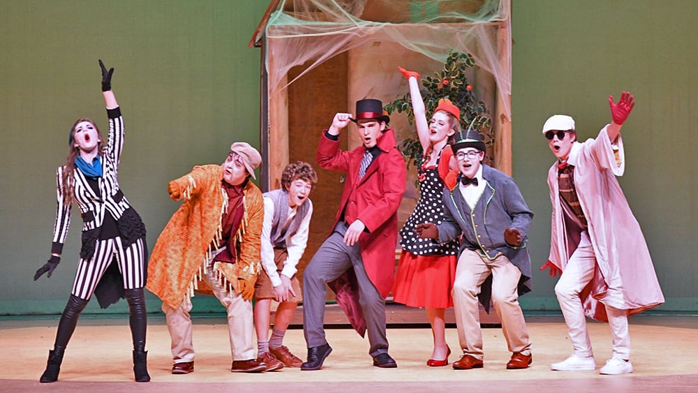 A group of performers in colorful, theatrical costumes stand on a stage against a green backdrop, striking dynamic poses and gestures.