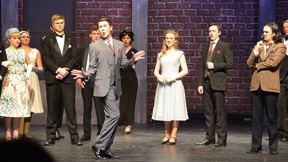 A group of formally dressed individuals, including a woman in a white dress, standing in front of a brick wall backdrop.