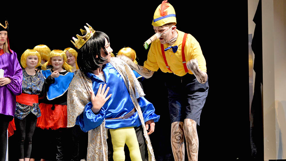 A group of colorfully dressed performers, including a person in a yellow costume with a hat, interacting on a stage with a dark background.