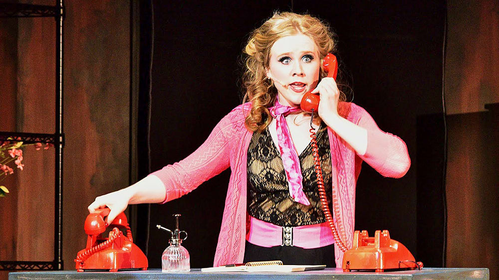 A woman with curly blonde hair wearing a bright pink outfit stands behind a table with two vintage-style telephones, appearing surprised or excited.