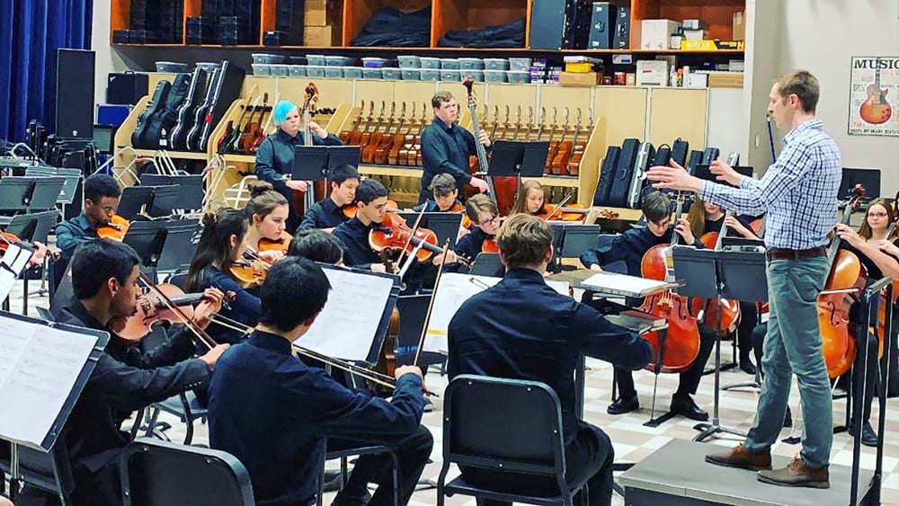 A group of musicians, including violinists, cellists, and a conductor, are performing on a stage in a music store filled with various musical instruments and equipment.