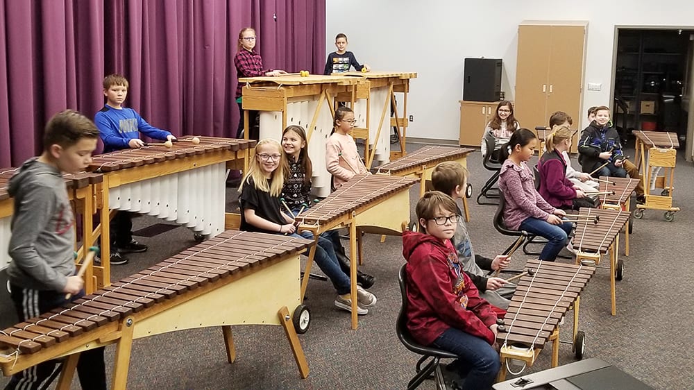 A group of people, mostly young students, are seated at various wooden instruments, including xylophones and marimbas, in what appears to be a music classroom or performance space with purple curtains in the background.