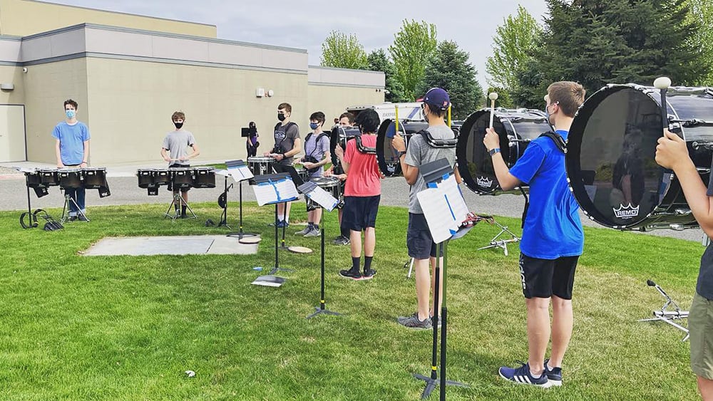A group of people, likely a musical ensemble, performing outdoors on a grassy field with a building in the background.