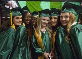 A group of young women in green graduation gowns and caps stand together, smiling and celebrating their academic achievement against a backdrop of green and black decorations.