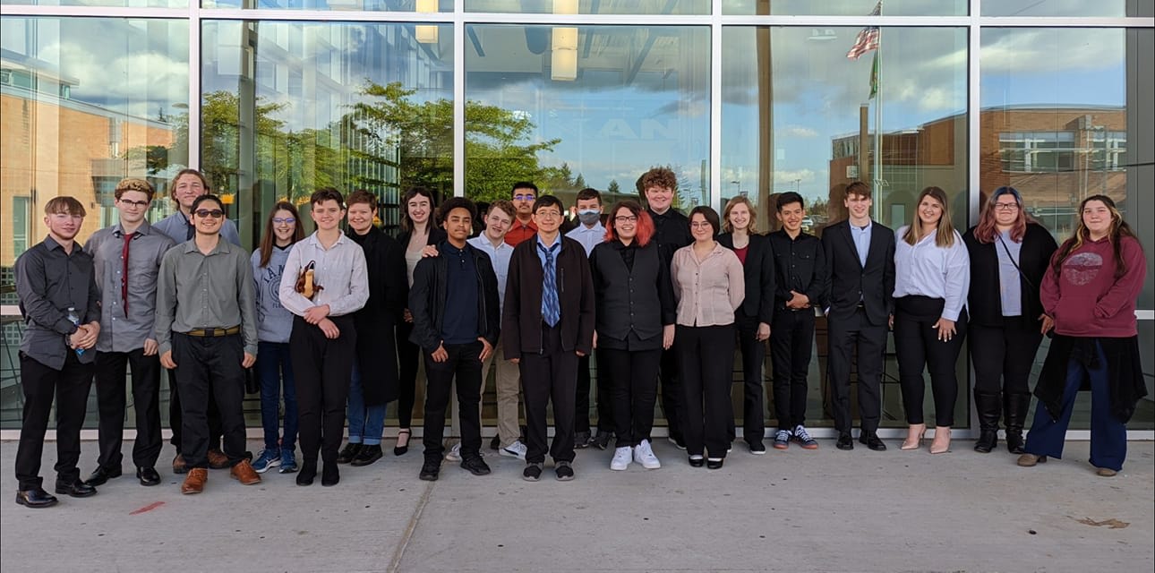 A large group of people, both men and women, standing together in front of a glass building with trees and a cloudy sky visible in the background.