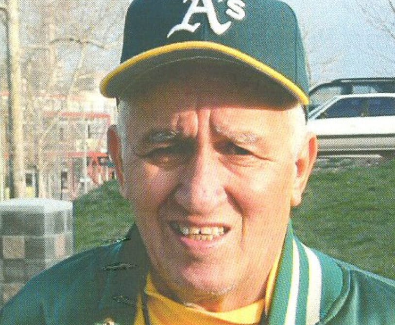 A photo of a smiling man in a green and yellow varsity jacket and cap.