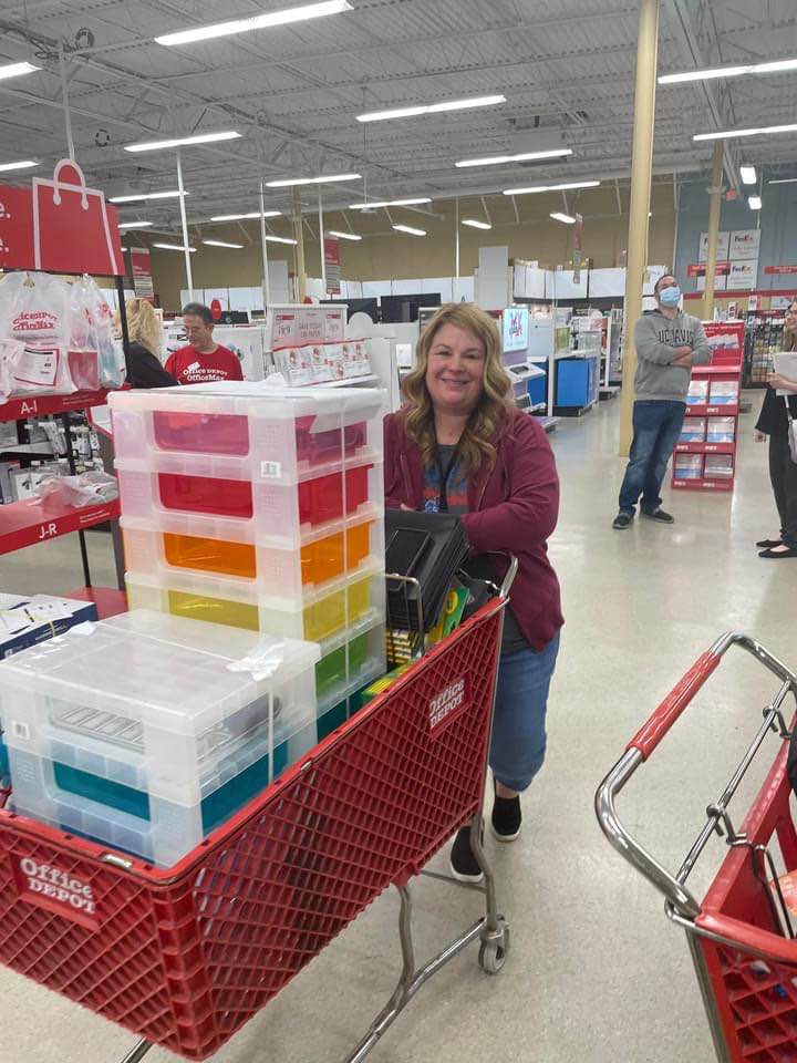 photo of a smiling teacher in a office supply store. she has a rainbow plastic drawer set in a red cart.
