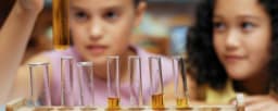 The image shows two young girls, one with blonde hair and the other with dark hair, looking intently at a row of test tubes filled with a yellow liquid.