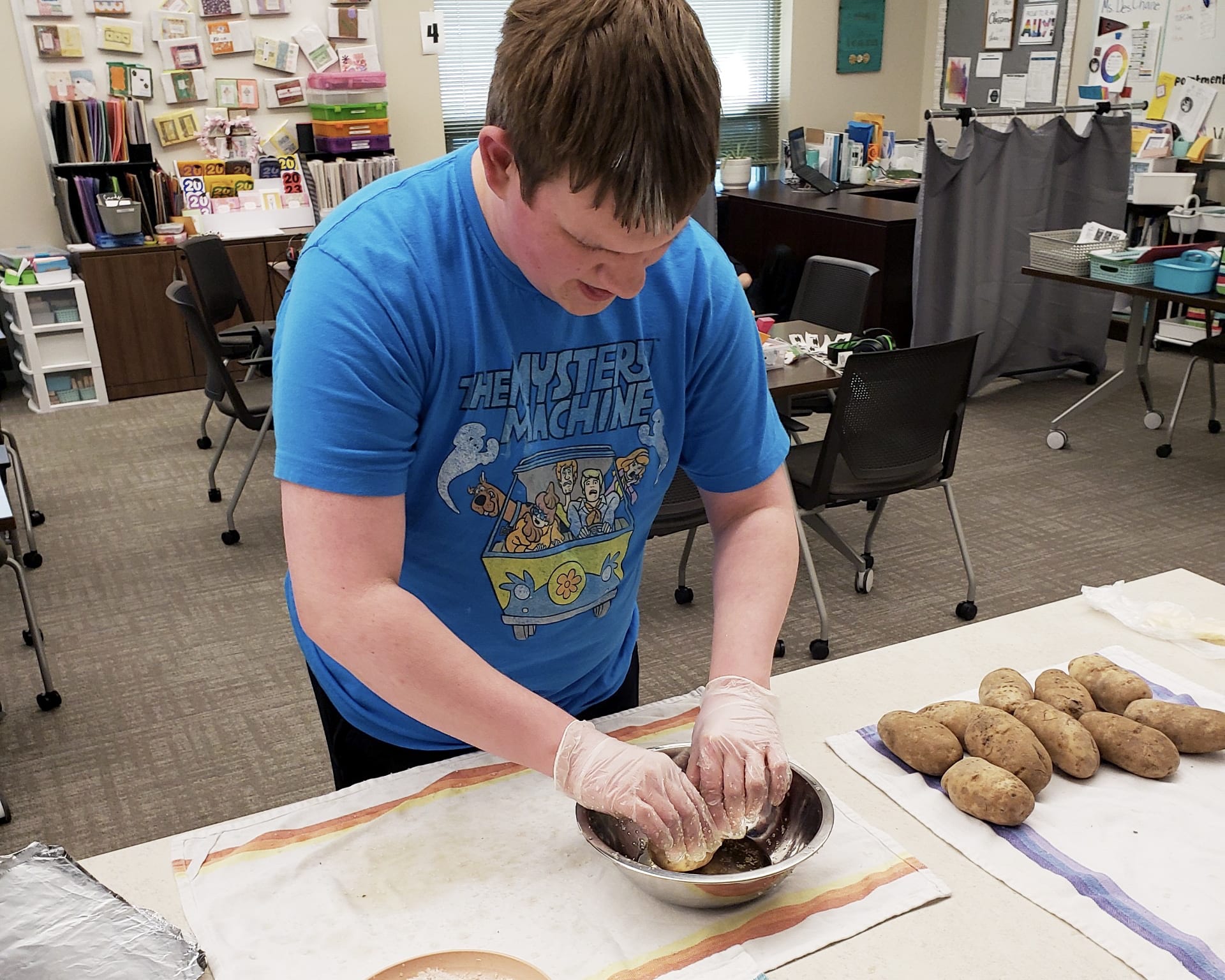 A young person in a blue t-shirt is working on a task at a desk in a classroom-like setting, with various items and materials visible in the background.