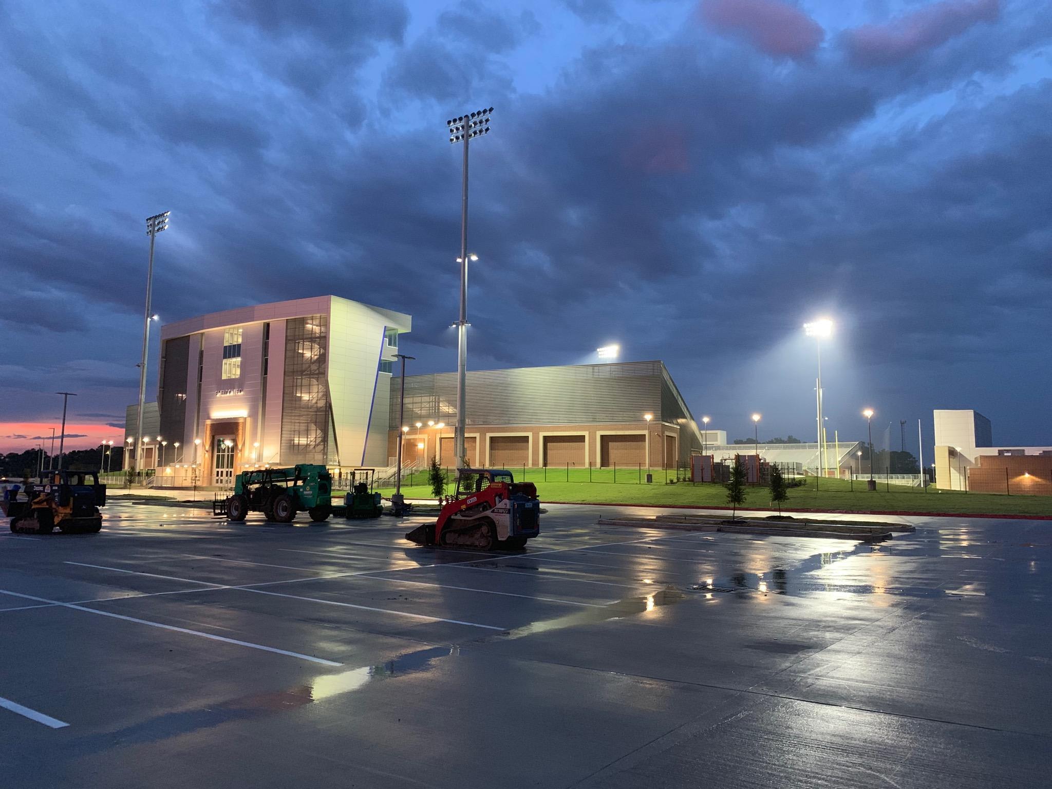 Construction equipment sits on a wet parking lot in front of a modern building under a cloudy twilight sky.