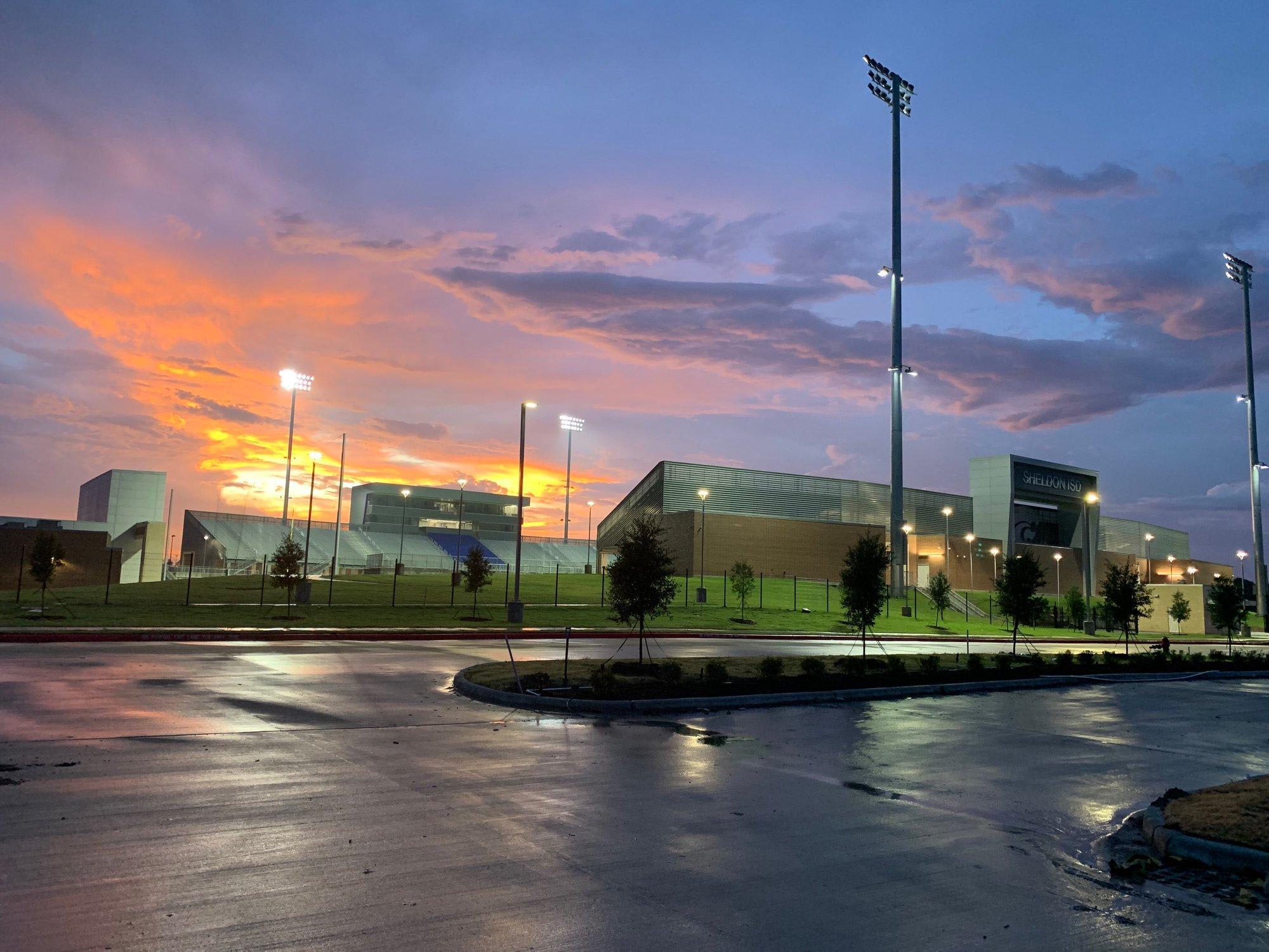 A wet asphalt parking lot sits in the foreground before a complex of modern buildings under a vibrant orange and purple sunset sky.