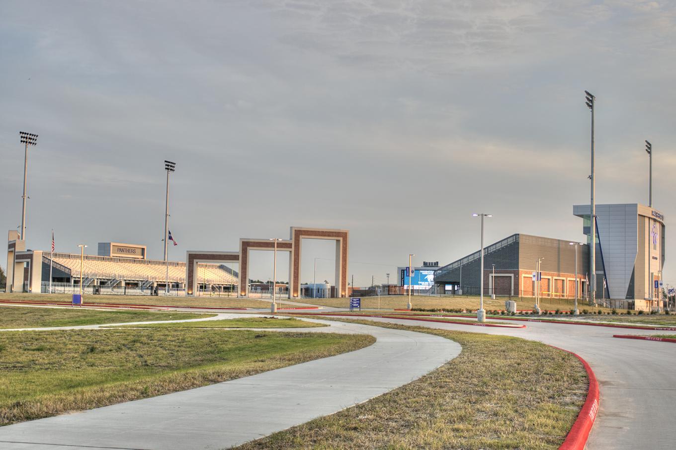 A curved concrete path and grassy area lead toward a complex of modern buildings and tall light poles under a pale sky.