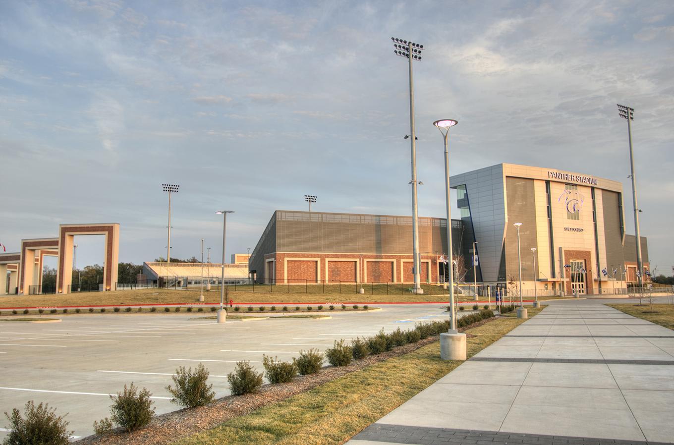 A concrete walkway and parking lot lead toward a large modern sports complex under a pale blue sky.
