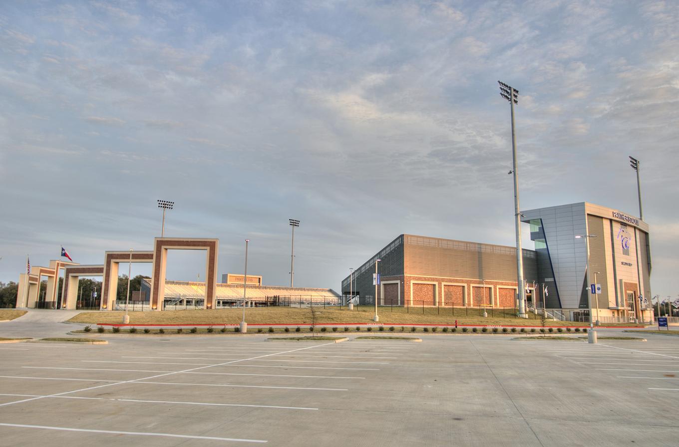 An empty concrete parking lot sits in the foreground before a complex of modern buildings and tall light poles under a cloudy sky.