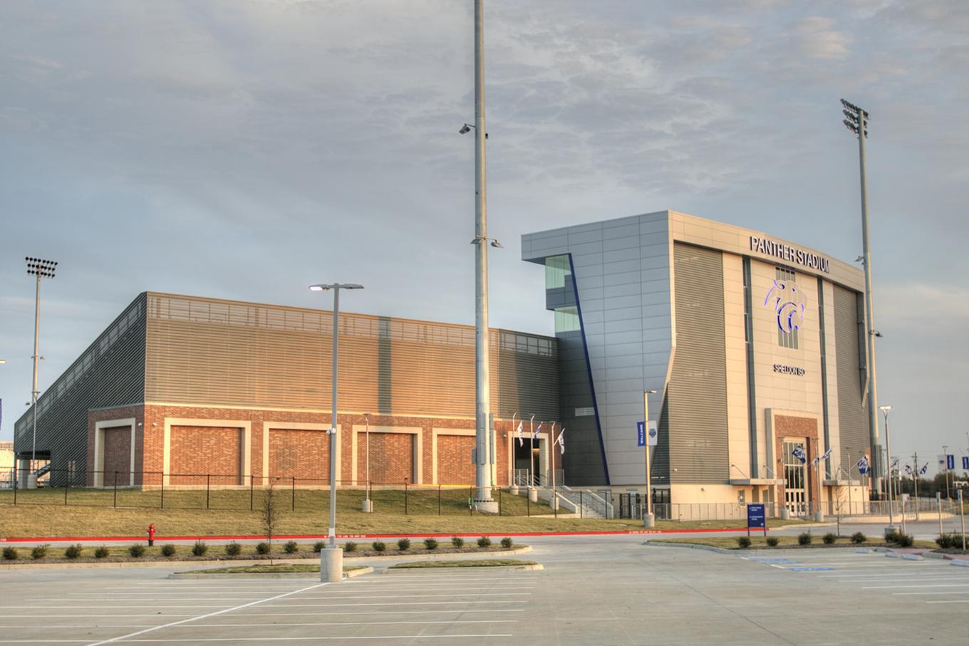 A large modern sports complex stands behind a concrete parking lot under a cloudy sky.