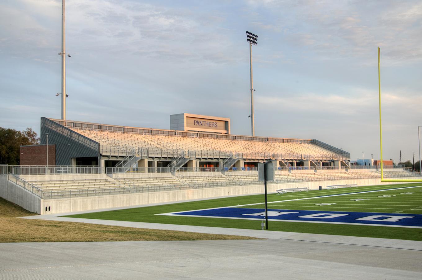 A green football field with white markings sits in the foreground, while a large stadium grandstand and tall light poles stand against a cloudy sky in the background.