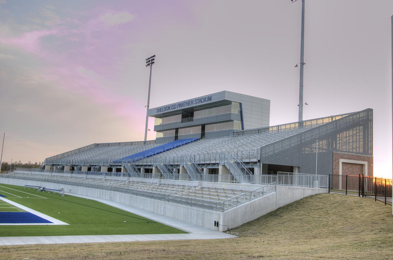 Empty bleachers and a green sports field sit in the foreground beneath a modern stadium building and a purple-hued sky.
