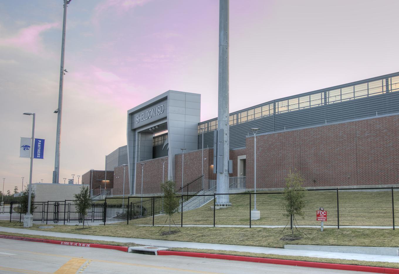 A paved road with a red curb sits in the foreground before a large brick and metal building under a pale purple sky.