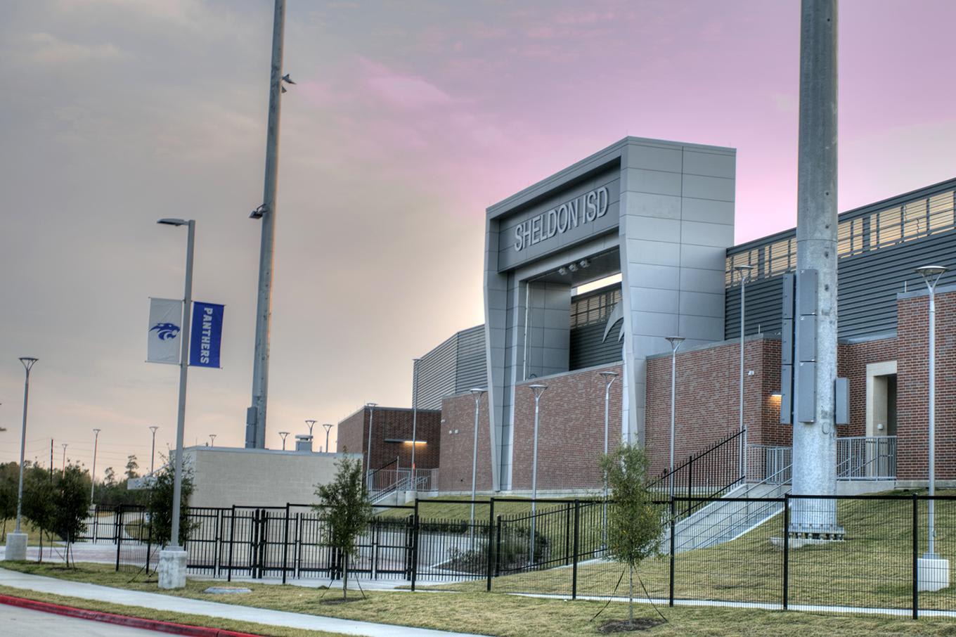 A modern school building labeled %22SHELDON ISD%22 stands behind a black fence and grassy area under a purple-tinted sky.