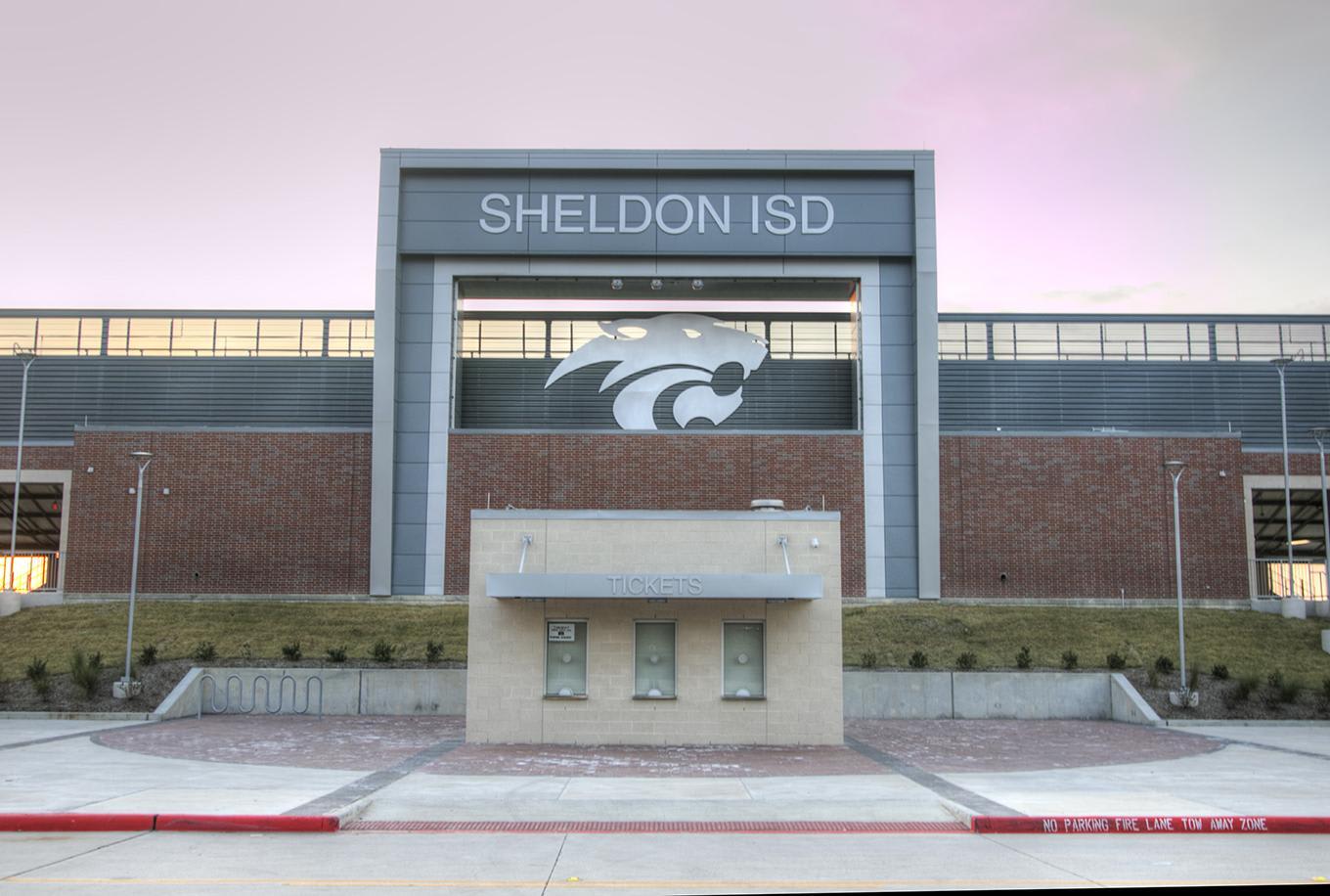 A concrete structure with three doors sits in the foreground before a large building labeled %22SHELDON ISD%22 under a pale sky.