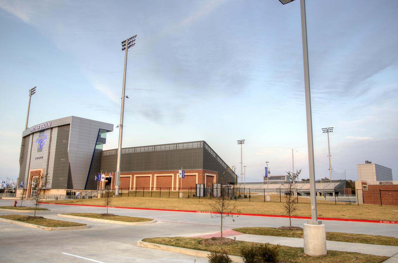 A modern sports stadium with tall light poles stands behind a paved parking area and grassy verge under a pale blue sky.