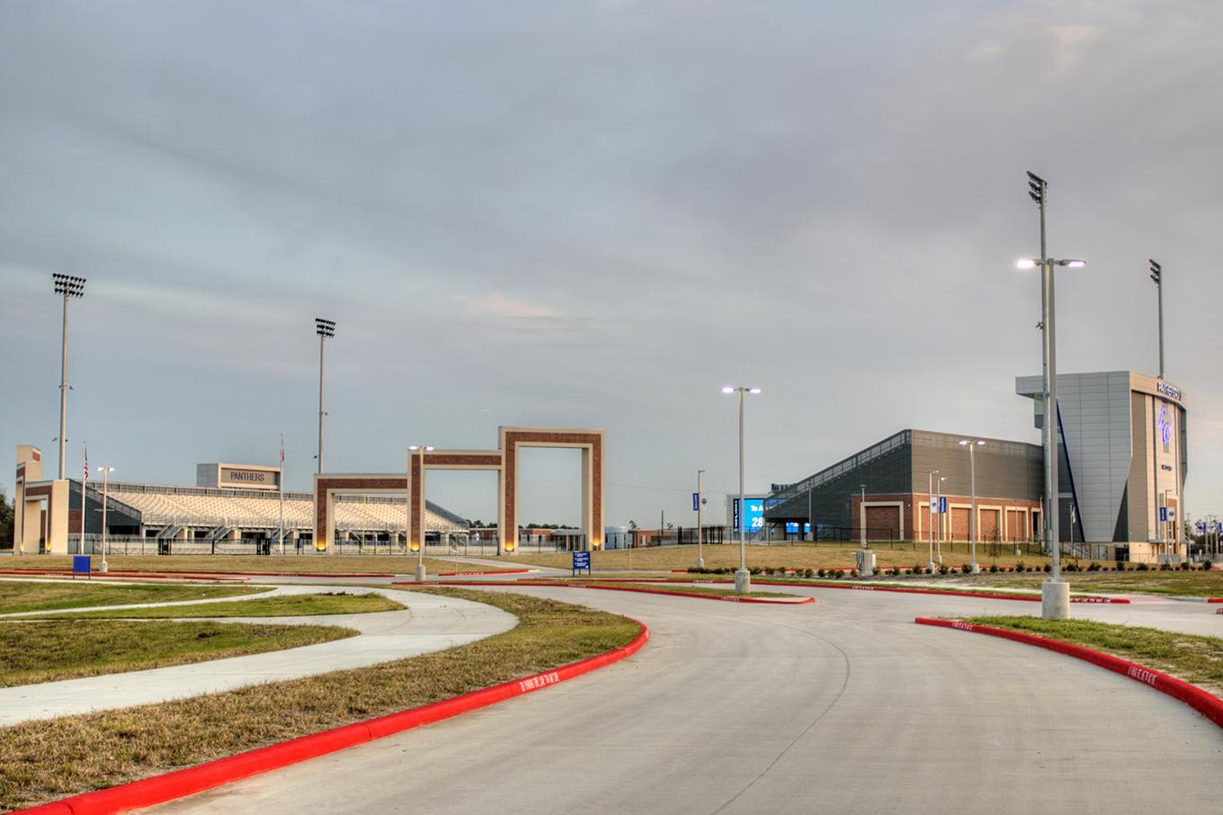 A curved concrete road with red curbs leads toward a modern sports complex and stadium under a cloudy sky.