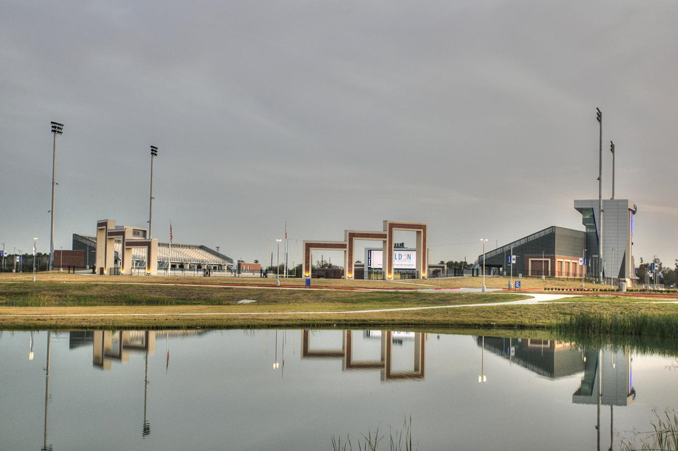 A calm pond in the foreground reflects a modern sports complex with large architectural frames and stadium lighting under a cloudy sky.