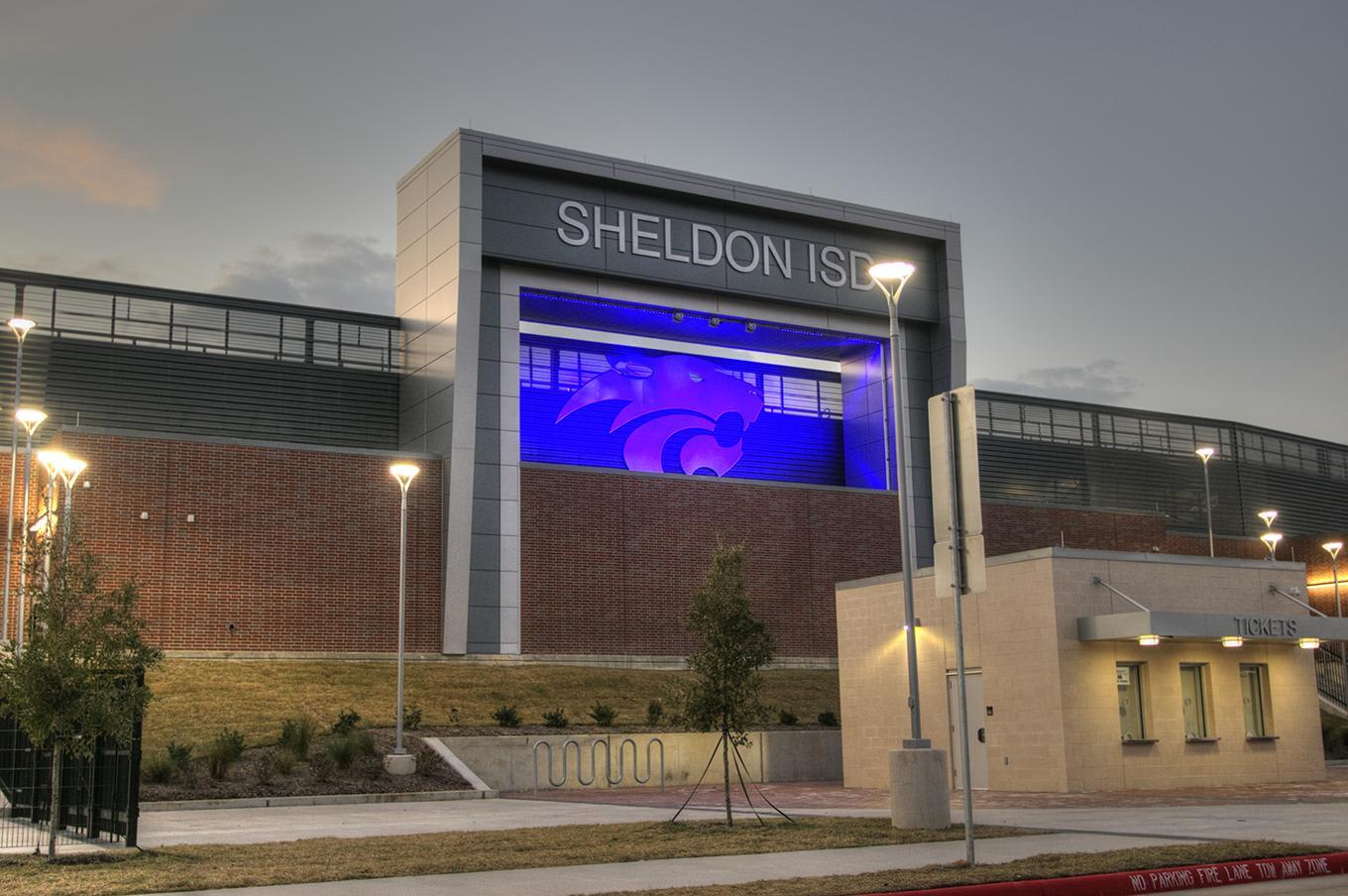 A modern school building with a large %22SHELDON ISD%22 sign and a purple logo stands under a dim sky, with a small utility building and young trees in the foreground.