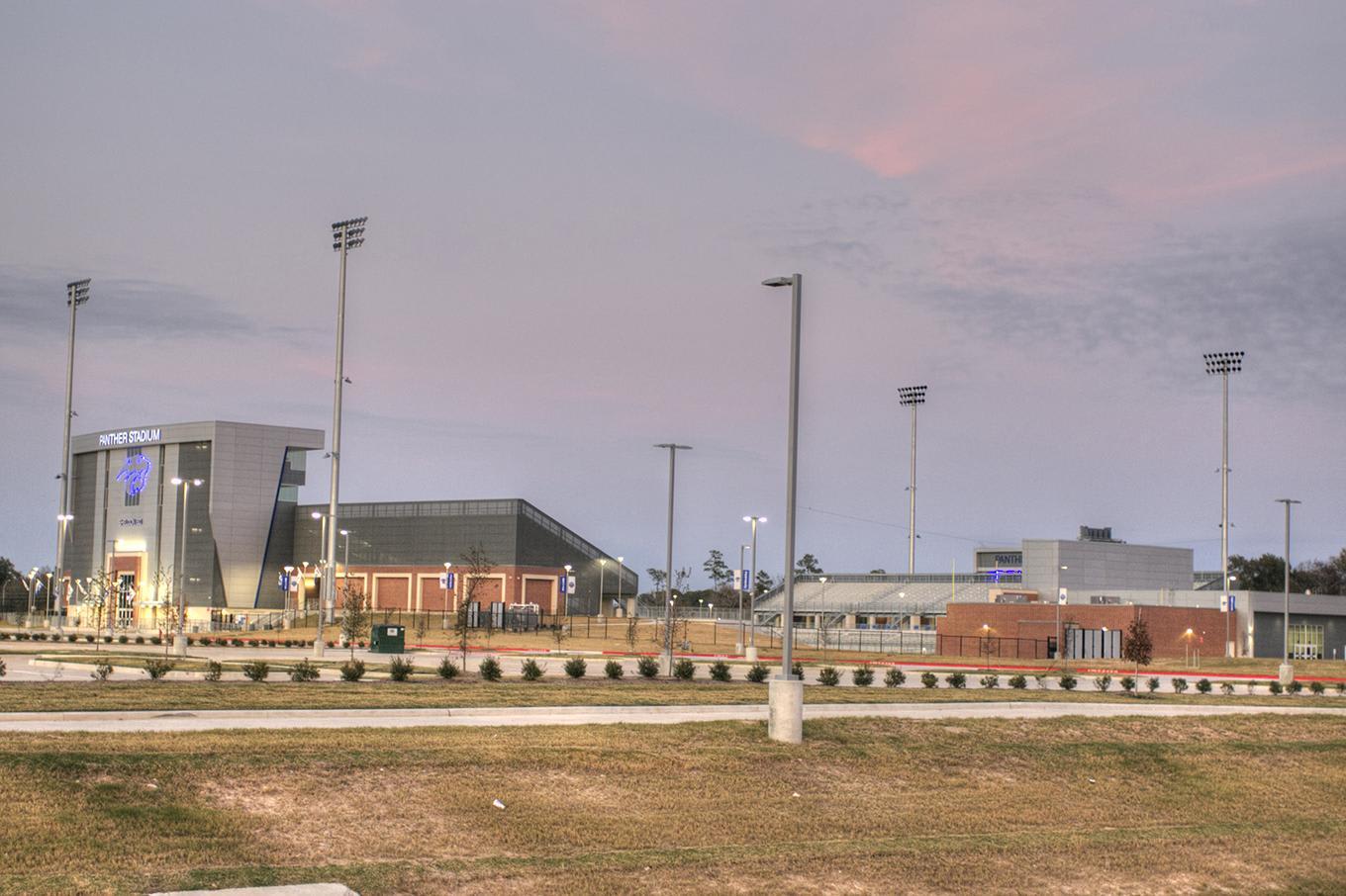 A grassy field and paved path lead toward a complex of modern sports facilities and tall light poles under a cloudy, sunset sky.
