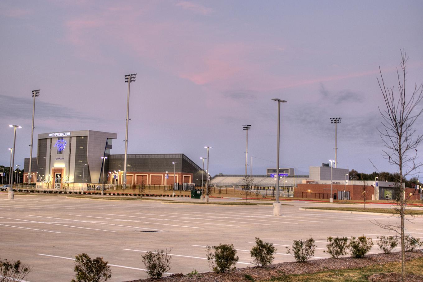 An empty parking lot sits in the foreground before a complex of modern buildings and tall light poles under a purple-hued twilight sky.