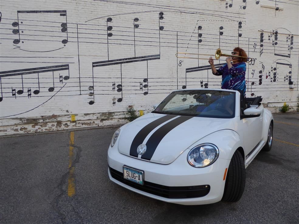 Anita McLaughlin playing the trombone while seated on the front seat of a white Volkswagen Beetle with black racing stripes on the hood. The car is parked in front of a white brick wall decorated with black music notes.