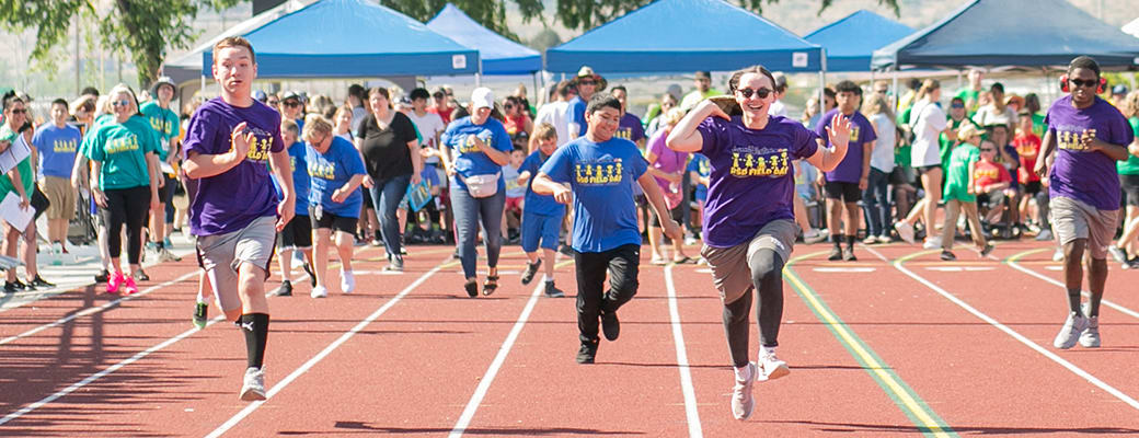 A group of people wearing purple shirts are running on a track surrounded by tents and a crowd of spectators.
